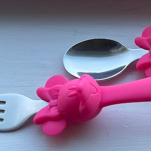 May include: Two pink children's spoons with a cartoon character handle. The spoons are made of stainless steel and have a pink plastic handle with a cartoon character design.