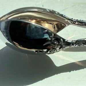 Puede incluir: Pinzas de servir de plata con un diseño ornamentado y un extremo en forma de cuchara. Las pinzas están cerradas y tienen un detalle decorativo en el centro.