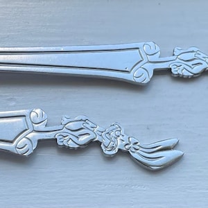 Puede incluir: Dos cucharas de plata con un diseño de conejo de dibujos animados en los mangos. El conejo está en una pose de carrera con las orejas y la cola extendidas.