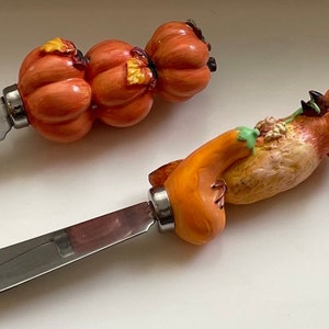 Peut inclure: Deux couteaux à beurre décoratifs. L'un présente une poignée avec trois citrouilles oranges, l'autre une figurine d'oiseau tenant une citrouille. Les deux couteaux ont des lames en acier inoxydable et sont conçus pour tartiner.