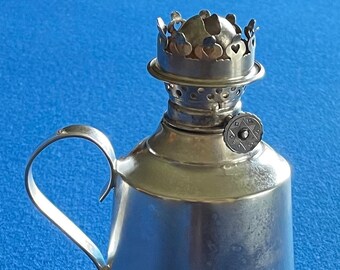 May include: A silver oil lamp with a handle and a decorative top. The lamp has a round base and a small, round knob on the side.