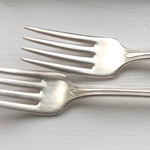 May include: Two silver forks with ornate handles. The forks are laid out side by side, with the tines pointing in opposite directions.