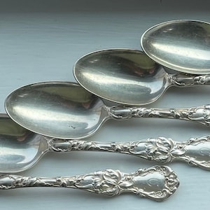 May include: Five silver teaspoons with ornate handles. The spoons are arranged in a staggered pattern, with the handles pointing to the right.