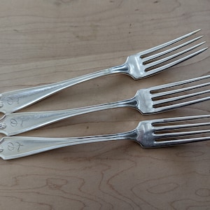 May include: Three silver forks with four tines each, arranged on a light wood surface. Each fork has a decorative handle end with a floral design and the engraved letters "07". These forks are suitable for a formal dining setting.