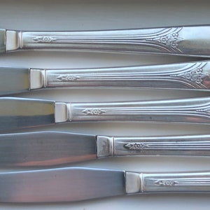 Op de afbeelding: Vijf zilveren botermesjes met sierlijke handvatten. De mesjes zijn in een rij gerangschikt, met de handvatten naar rechts wijzend.