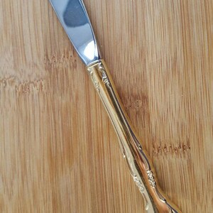 May include: A silver butter knife with a gold handle featuring an ornate floral design. The knife is on a wooden surface.