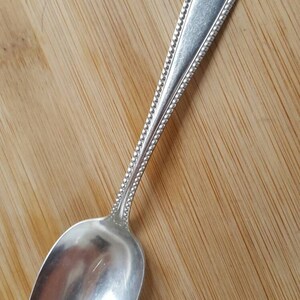 May include: A silver teaspoon with a beaded handle and a simple, rounded bowl. The spoon is lying on a wooden surface.