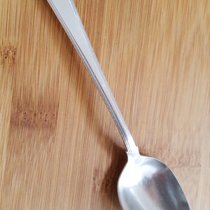 May include: A silver teaspoon with an ornate handle and the inscription "Grace" on the front. The spoon is lying on a wooden surface.