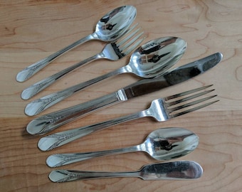 May include: A set of six silver-plated flatware pieces, including a butter knife, a teaspoon, a fork, and three spoons. The flatware is arranged on a wooden surface.