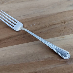 May include: A silver fork with four tines, resting on a wooden surface. The handle features an ornate floral design. This vintage utensil is likely part of a formal dining set.