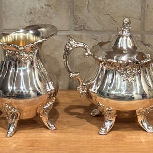 May include: A silver creamer and sugar bowl set with ornate detailing and three legs each. The creamer has a handle and a spout. The sugar bowl has a lid.