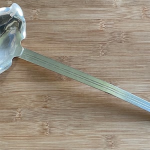 May include: A silver ladle with a decorative, scalloped bowl and a long handle. The handle features three parallel grooves. The ladle is made of a shiny, reflective metal, likely stainless steel, and is designed for serving soups or sauces.