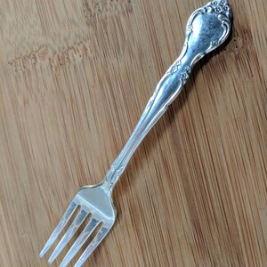 May include: A silver fork with an ornate handle and a decorative design. The fork is on a wooden surface.