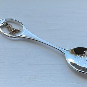 May include: Silver souvenir spoon with a charm of a boot hanging from the handle. The spoon has an image of a building with the word "Dallas" on it.