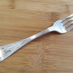 May include: A silver fork with an ornate handle. The fork has four tines and is lying on a wooden surface.