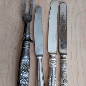 May include: Four silver utensils, including a fork and three butter knives, are arranged on a light wood surface. The fork has a decorative handle with a floral design. The butter knives have ornate handles with floral designs. One of the butter knives has the initials "NR" engraved on the blade.