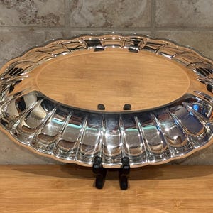 Può includere: Un vassoio ovale color argento con bordo smerlato e interno in legno. Il vassoio è sostenuto da un supporto nero. La superficie riflettente del vassoio cattura la luce, evidenziando il suo design ornato.