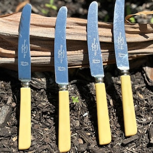 May include: Four vintage knives with stainless steel blades and cream-coloured handles. The blades are curved and stamped with text. The handles are a light yellow colour. The knives are arranged on dark mulch.