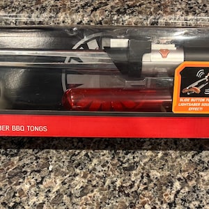 Peut inclure: Un ensemble de pinces à barbecue Star Wars Darth Vader Lightsaber dans un emballage en plastique transparent. Les pinces sont conçues pour ressembler à un sabre laser, avec une poignée rouge et une lame bleue éclairée. L'emballage présente le logo Star Wars et une image de Dark Vador.
