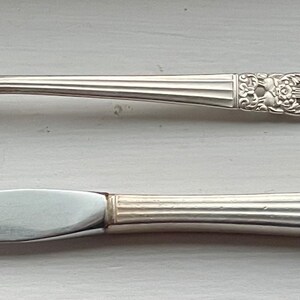 Könnte beinhalten: Eine silberne Gabel und ein Messer mit verzierten Griffen. Die Griffe haben ein dekoratives Design mit einem floralen Motiv.