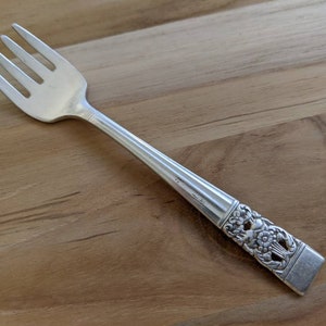May include: A silver fork with an ornate design on the handle. The fork has four tines and a rounded handle.