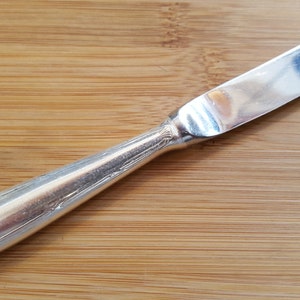 May include: A silver butter knife with an ornate handle. The knife is on a wooden surface.