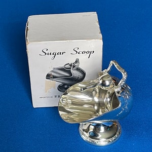 Op de afbeelding: Een zilveren suikerlepel met een sierlijk ontwerp en een handvat. De lepel ligt op een witte doos met de woorden "Sugar Scoop" erop gedrukt.
