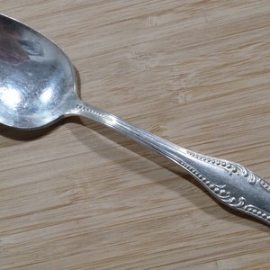 Op de afbeelding: Een zilveren lepel met een sierlijk handvat. De lepel heeft een ronde kom en een decoratief ontwerp op het handvat.