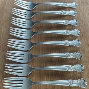 May include: Set of eight silver forks with ornate handles featuring a floral design. Each fork has a letter 'S' engraved on the handle.