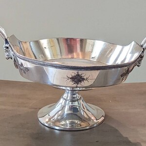 Può includere: Una zuppiera d'argento con due manici e una base a piedistallo. La zuppiera ha un design decorativo sul fondo.