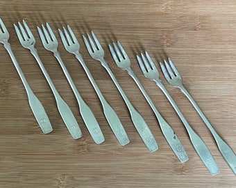 Op de afbeelding: Acht zilveren vorken met een eenvoudig ontwerp, in een rij op een houten oppervlak gelegd. De vorken zijn allemaal even groot en hebben dezelfde vorm, en ze hebben een licht gebogen handvat.