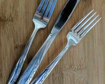 May include: Three pieces of silverware, a knife and two forks, with ornate handles, on a wooden surface.