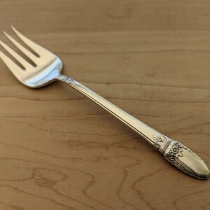 May include: A silver fork with an ornate handle. The handle features a floral design and is slightly curved. The fork is resting on a wooden surface.