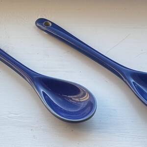 May include: Two navy blue ceramic spoons with a glossy finish are displayed. Each spoon has a long handle with a small hole at the end for hanging. The spoons are designed for serving or cooking.