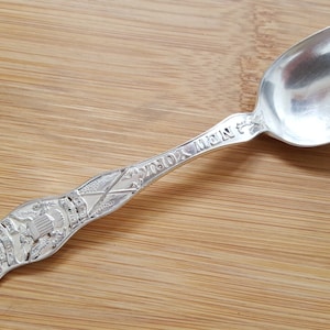 May include: A silver spoon with an ornate design featuring a building and the words "NEW YORK" on the handle.