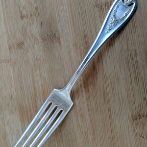 May include: A silver fork with a decorative handle featuring floral and leaf designs. The fork has four tines and is set against a wooden background. The handle has a detailed, ornate pattern.