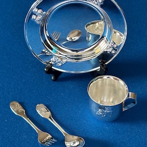 May include: A silver baby set with a plate, cup, spoon, and fork. The plate and cup have grape designs. The spoon and fork have a baby design.