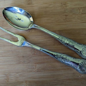 Könnte beinhalten: Ein silbernes Servierlöffel- und Gabel-Set. Der Löffel hat eine große ovale Schüssel, und die Gabel hat zwei Zinken. Beide Utensilien haben verzierte Blumen- und Muscheldekore an den Griffen.