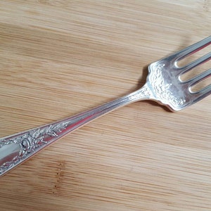Könnte beinhalten: Eine silberne Gabel mit einem verzierten Design am Griff. Die Gabel hat vier Zinken und liegt auf einer Holzoberfläche.