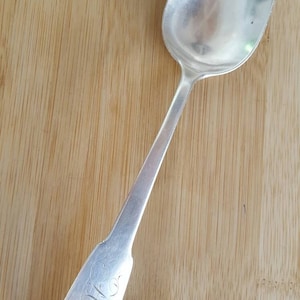May include: A silver spoon with an ornate handle featuring a shell design. The spoon has a long, slender handle and a rounded bowl.