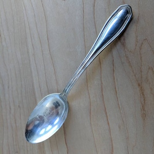 May include: A silver teaspoon with a rounded bowl and a simple, elegant design. The spoon is lying on a light wood surface.