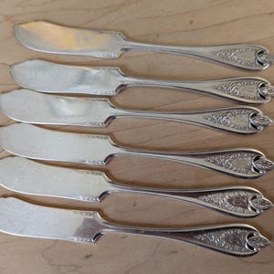 May include: Set of six silver butter knives with ornate handles. The knives have a polished finish and are arranged in a row.
