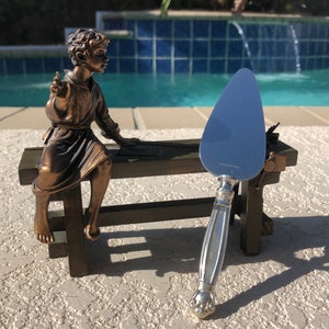 May include: A silver cake server with an ornate handle and a pointed blade. The server is resting on a wooden bench with a bronze statue of a boy sitting on it.