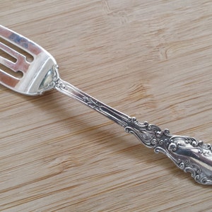 May include: A silver fork with three tines and an ornate handle. The fork is made of a shiny metal and has a decorative floral pattern on the handle. The fork is resting on a light brown wooden surface.
