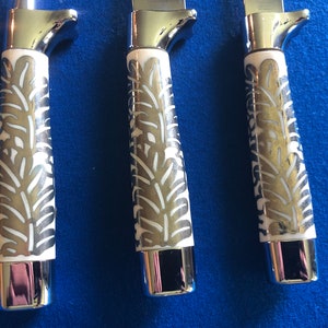 May include: Three silver knives with white handles featuring a decorative leaf pattern. The handles are made of a white material with a silver leaf design.