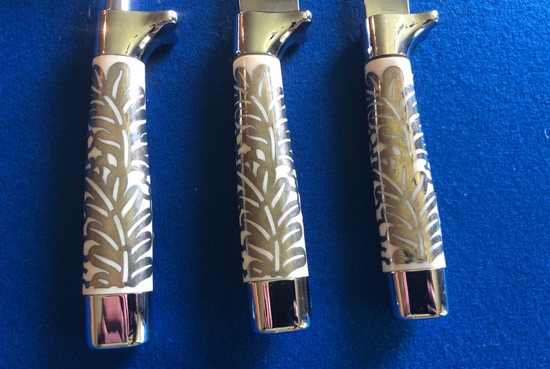 May include: Three silver knives with white handles featuring a decorative leaf pattern. The handles are made of a white material with a silver leaf design.