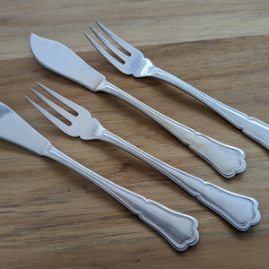 May include: Four silver-toned utensils, two butter knives and two forks, with ornate handles, laid out on a wooden surface.