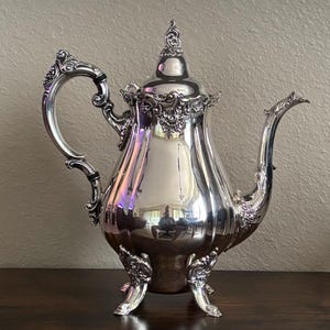 May include: A polished silver-plated coffee pot featuring an ornate, scrolled handle and a matching curved spout. The body is smooth and reflective, accented with intricate floral relief detailing around the base, lid, and handle attachment points.