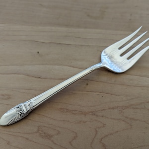 May include: A silver fork with an ornate handle and four prongs. The fork is on a wooden surface.