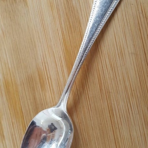 May include: A silver teaspoon with a beaded handle. The spoon is on a wooden surface.